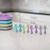 Bumkins Silicone Chewtensils®: Lavender are arranged with other silicone utensils near a stack of divided plates on a marble kitchen counter