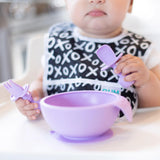 Close-up of a baby in a high chair, wearing a black & white bib as they eat from a purple bowl with Bumkins Silicone Chewtensils®: Lavender.