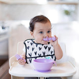 A toddler wearing a heart-patterned bib in a high chair eats from a purple bowl using Bumkins Silicone Chewtensils®: Lavender.