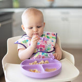 A curious baby in a colorful bib sits in a high chair, eating from Bumkins Grip Dish Essential Infant 4-Piece Mealtime Set: Lavender.