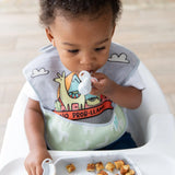 A toddler in a high chair, wearing a llama bib, holds a Bumkins Silicone Chewtensils®: Marble in their mouth; a plate of food sits in front.