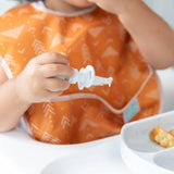 Close-up of a toddler in an orange bib gripping a Bumkins Silicone Chewtensils®: Marble with a matching plate of food nearby.