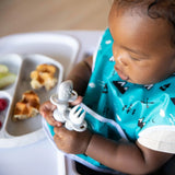 A toddler in a teal bib holds Bumkins Silicone Chewtensils®: Marble near a plate with raspberries, waffle pieces, and apple slices.