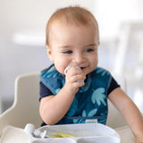 A baby in a high chair grins, wearing a blue bib and chewing a Bumkins Silicone Chewtensils®: Marble.