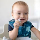 A smiling baby in a high chair wearing a blue bib chews a Bumkins Silicone Chewtensils®: Marble.