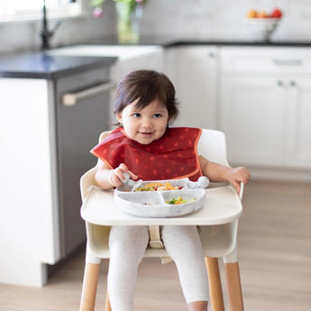 A toddler in a high chair wearing a red bib smiles as they use Bumkins Silicone Chewtensils®: Marble to eat from a matching plate.