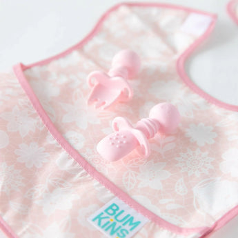 Bumkins Silicone Chewtensils®: Pink lay on top of a pink bib on a white background; one lays face down, showing sensory bumps on the back.