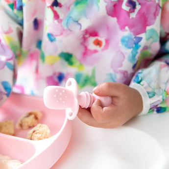 Close-up of a child in a colorful bib holding a Bumkins Silicone Chewtensils®: Pink next to a pink divided plate.