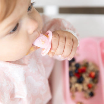 Close-up of a baby in a pink bib with a Bumkins Silicone Chewtensils®: Pink, enjoying baby-led weaning with oats & berries on a pink plate.