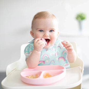 A baby in a high chair wearing a rainbow-print bib enjoys baby-led weaning from a pink plate with Bumkins Silicone Chewtensils®: Pink.