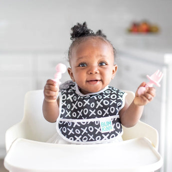 A smiling baby in a high chair wearing a black & white bib holds Bumkins Silicone Chewtensils®: Pink.