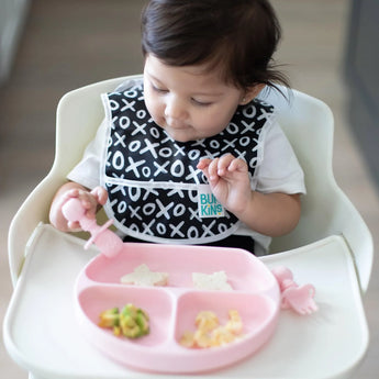A baby in a high chair wearing a black & white bib enjoys a meal from a pink divided plate with Bumkins Silicone Chewtensils®: Pink.