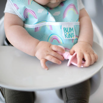 A baby in a high chair wearing a rainbow bib grasps a Bumkins Silicone Chewtensils®: Pink.