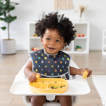 A toddler sits in a high chair, wearing a black patterned bib and eating from a yellow plate with Bumkins Silicone Chewtensils®: Pineapple.