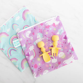Two colorful unicorn and rainbow patterned bags lay on a marble background, with Bumkins Silicone Chewtensils®: Pineapple on top.
