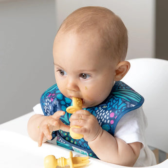 A baby in a high chair wearing a blue animal-print bib chews Bumkins Silicone Chewtensils®: Pineapple.