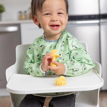 A happy toddler in a high chair, wearing a green bib, holds Bumkins Silicone Chewtensils®: Pineapple in a modern kitchen.