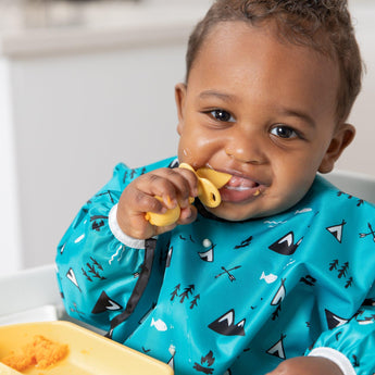 A baby in a high chair smiles with a Bumkins Silicone Chewtensils®: Pineapple near their mouth, wearing a blue patterned bib.