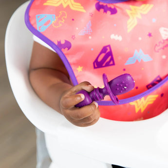 Close-up of a baby in a high chair holding a Bumkins Silicone Chewtensils®: Purple, wearing a colorful superhero-themed bib.