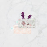 Bumkins Silicone Chewtensils®: Purple peeks from a floral zippered pouch, with another pink pouch underneath on a marble background.