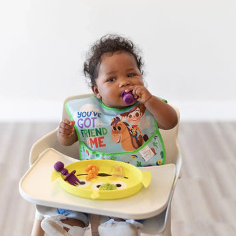 A child in a bib sits in a high chair, holding a Bumkins Silicone Chewtensils®: Purple in their mouth with a plate of food on the tray.