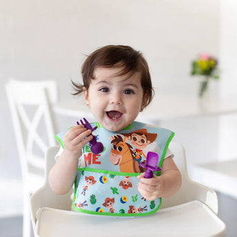 A smiling toddler in a high chair, wearing a colorful bib, holds Bumkins Silicone Chewtensils®: Purple in a bright room.
