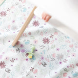 A Bumkins Silicone Chewtensils®: Sage with applesauce lays on a floral mat underneath a high chair with a baby's foot visible.