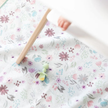 A Bumkins Silicone Chewtensils®: Sage with applesauce lays on a floral mat underneath a high chair with a baby's foot visible.