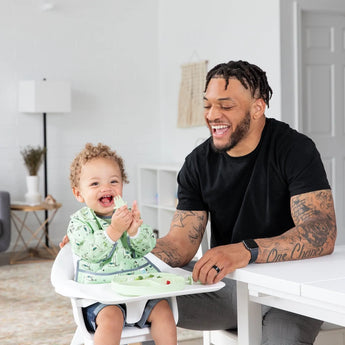 A man smiles at a table as a curly-haired child in a high chair enjoys baby-led weaning from a plate with Bumkins Silicone Chewtensils®: Sage.