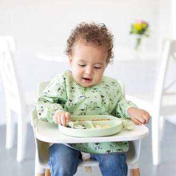 A toddler sits in a high chair, wearing a green bib and using Bumkins Silicone Chewtensils®: Sage to eat from a green divided plate.