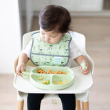 A toddler in a high chair wearing a green bib holds Bumkins Silicone Chewtensils®: Sage near a matching plate with food.