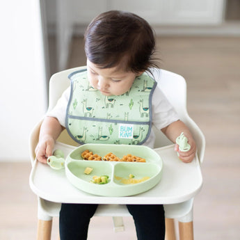 A toddler in a high chair wearing a green bib holds Bumkins Silicone Chewtensils®: Sage near a matching plate with food.