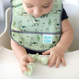 A baby in a high chair wearing a green bib plays with Bumkins Silicone Chewtensils®: Sage on the tray.