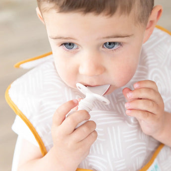 A toddler wearing a beige bib chews on a Bumkins Silicone Chewtensils®: Sand.