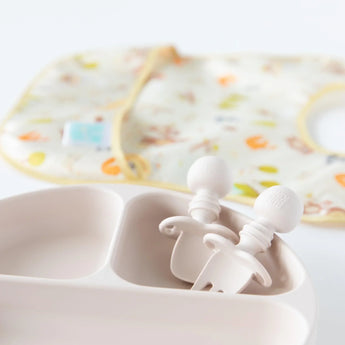 Close-up of Bumkins Silicone Chewtensils®: Sand in a section of a matching plate on a white background, with a blurred bib behind.