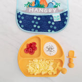 An orange plate with eggs, raspberries, and yogurt sits beside Bumkins Silicone Chewtensils®: Tangerine and a blue bib on a marble surface.