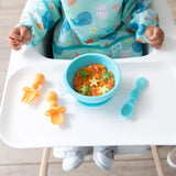 A child sits in a high chair wearing a blue bib, with a blue bowl of food, spoon, and Bumkins Silicone Chewtensils®: Tangerine on the tray.