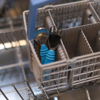 Bumkins Spoon + Fork: Dark Blue sits in the utensil compartment of an open dishwasher.