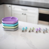 Bumkins Spoon + Fork: Lavender sits among other utensil sets and a stack of silicone divided plates on a marble kitchen counter.