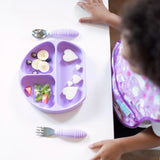 Top down view of a child at a white table with a purple divided plate of food and Bumkins Spoon + Fork: Lavender.