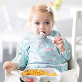 A toddler in a high chair eats pasta with Bumkins Spoon + Fork: Marble from a matching plate, wearing a bib featuring rainbow patterns.