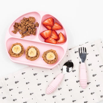 A pink plate with granola, strawberries, and bananas sits next to Bumkins Spoon + Fork: Pink on a patterned napkin on a white background.