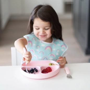 A young girl wearing a rainbow-patterned bib uses Bumkins Spoon + Fork: Pink to eat fruit from a pink divided plate at a white table.