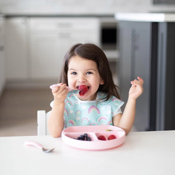 A child uses Bumkins Spoon + Fork: Pink to eat berries from a pink plate at a white table, smiling while wearing a rainbow-patterned bib.