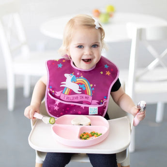 A toddler in a high chair wearing a unicorn bib smiles while holding Bumkins Spoon + Fork: Pink near a pink plate with a sandwich and veggies.