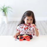 A child with long hair in a pink bib eats berries and pasta from a black & red divided plate with Bumkins Spoon + Fork: Red at a table.