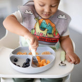 A toddler wearing a llama bib in a high chair uses Bumkins Spoon + Fork: Sage to eat fruit and waffles from a gray sectioned plate.