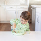 A child in a green sleeved bib uses Bumkins Spoon + Fork: Sage to eat from a green divided plate at a white table in a kitchen.