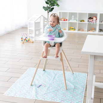 A toddler eats from a purple bowl in a high chair while Bumkins Splat Mat: Rainbows covers the floor below; a purple spoon sits on top.