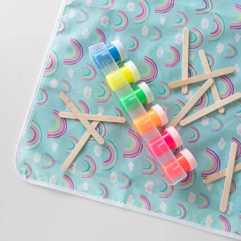Close-up of Bumkins Splat Mat: Rainbows on a white background, with colorful paint pots and wooden craft sticks on top.